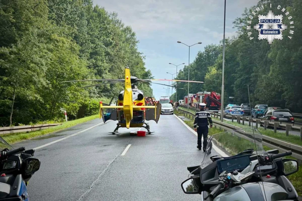 Śmigłowiec ratunkowy stojący na środku drogi ekspresowej, otoczony przez pojazdy policji i służb ratunkowych, policjant kieruje ruchem, na poboczu las i kolumna zatrzymanych samochodów.