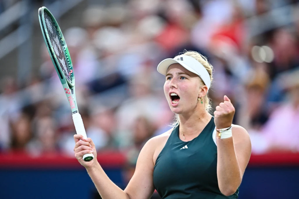 Tenisistka w ciemnozielonym stroju i białej opasce na głowie z wyraźną ekspresją triumfu, trzymająca rakietę w prawej ręce, zaciśnięta pięść w geście zwycięstwa, w tle rozmazana publiczność na trybunach.