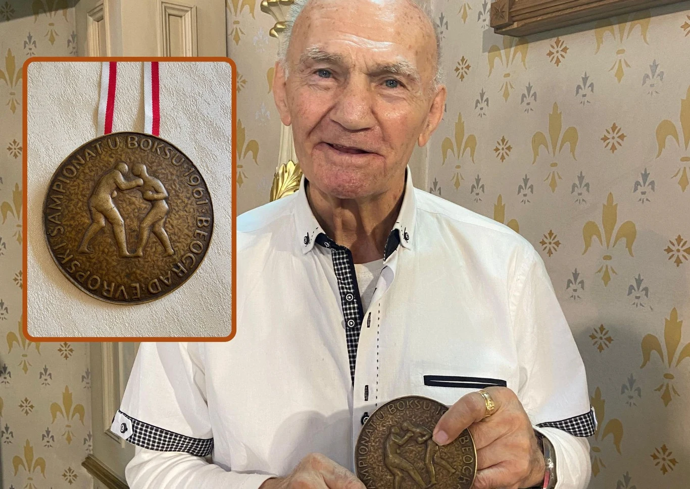 Starszy mężczyzna w białej koszuli trzyma w dłoni dużą medalionową nagrodę z motywem dwójki bokserów w walce oraz napisem 'MISTRZOSTWA EUROPY W BOKSIE'. W rogu zdjęcia znajduje się powiększenie tego medalu z zawieszką.