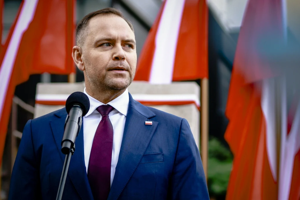 Mężczyzna w garniturze i bordowym krawacie stoi przy mikrofonie, w tle liczne biało-czerwone flagi, atmosfera oficjalna związana z uroczystością państwową.