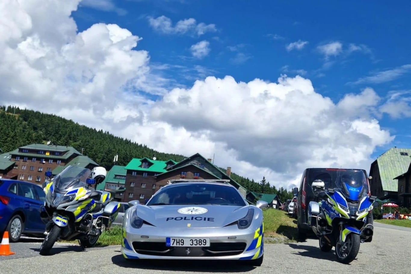 Radiowóz policyjny sportowy oraz dwa policyjne motocykle zaparkowane na tle budynków górskich, niebo z gęstymi chmurami, całość otoczona przyrodą i samochodami cywilnymi.