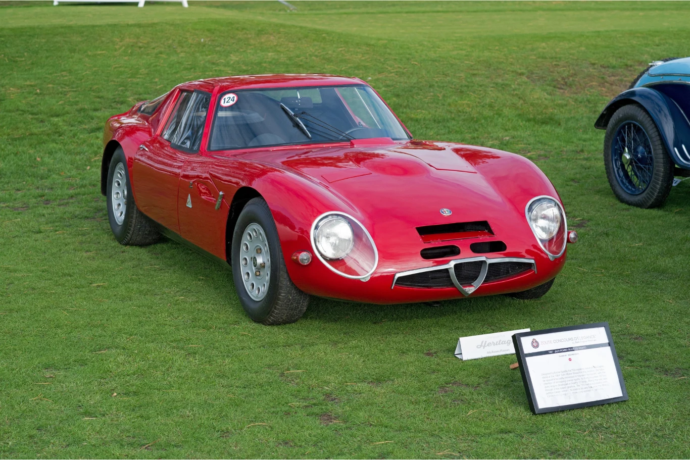 Czerwony klasyczny samochód sportowy z niskim nadwoziem ustawiony na trawie, przed pojazdem stoi tabliczka z informacjami o eksponacie.