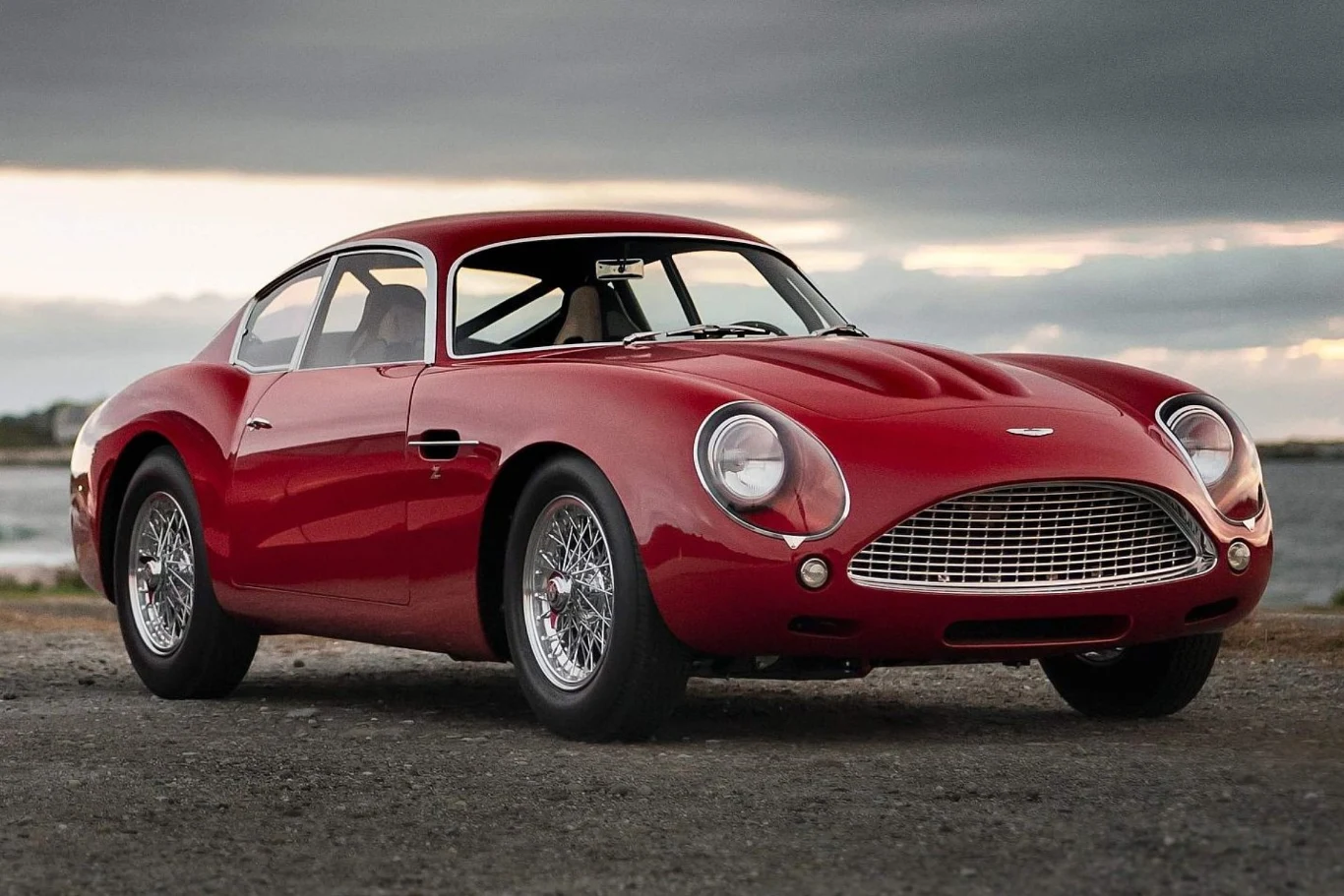 Czerwony klasyczny samochód sportowy Aston Martin DB4 GT Zagato ustawiony na tle pochmurnego nieba, ze szczegółowo widoczną przednią maską, chromowaną kratką oraz reflektorami.
