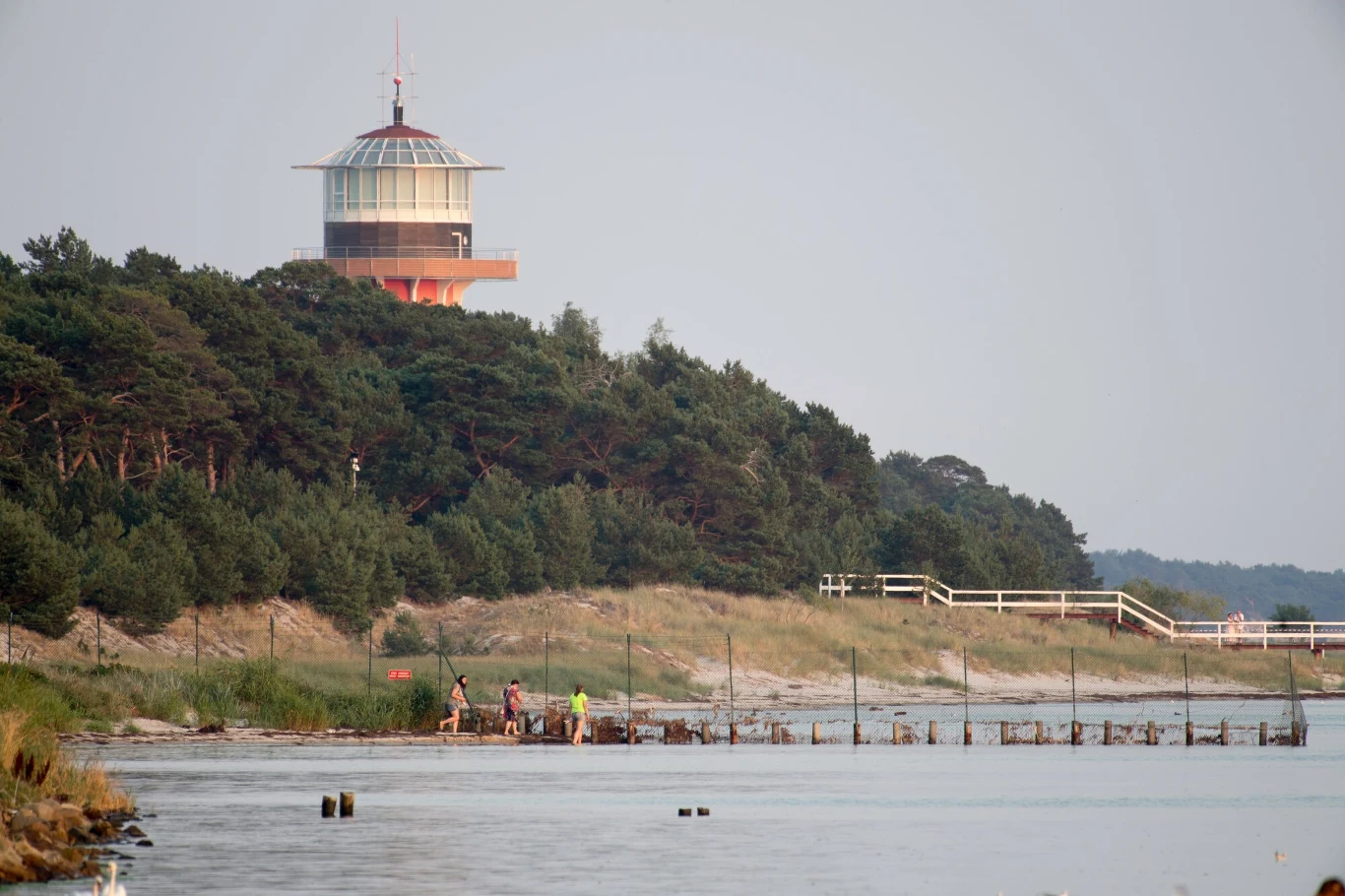 Rezydencja prezydenta RP w Juracie Wieża widokowa na tle zalesionego wzgórza, w dolnej części obrazu fragment linii brzegowej z piaskiem i trawą, kilka osób pracujących lub spacerujących przy wodzie, w tle widać drewniane pomosty i wały ochronne.