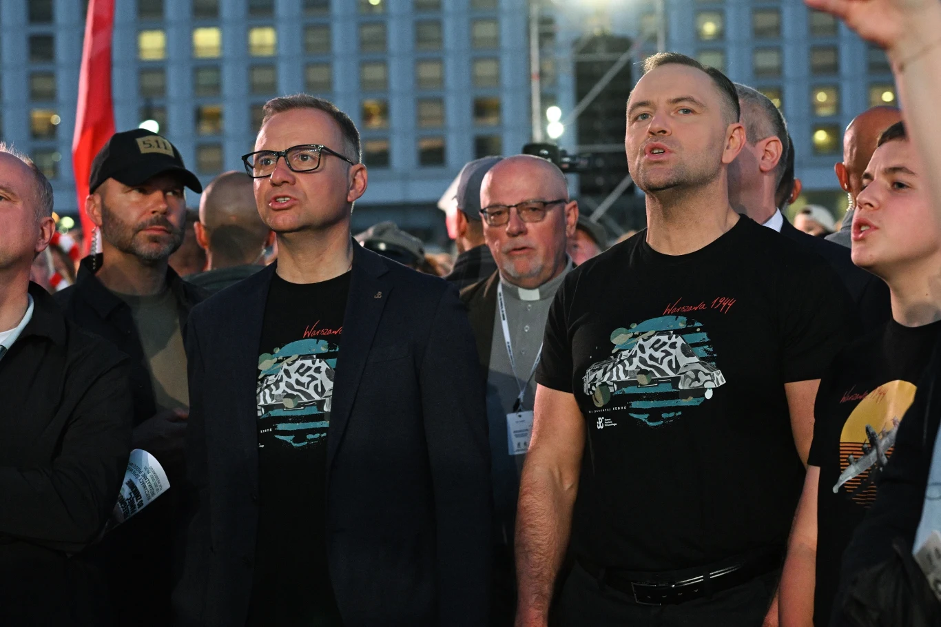 Andrzej Duda i Karol Nawrocki Grupa mężczyzn stojących razem na tle wysokiego budynku, część z nich ubrana w czarne koszulki z grafiką, jeden mężczyzna w koloratce, atmosfera wydarzenia społecznego lub publicznego.
