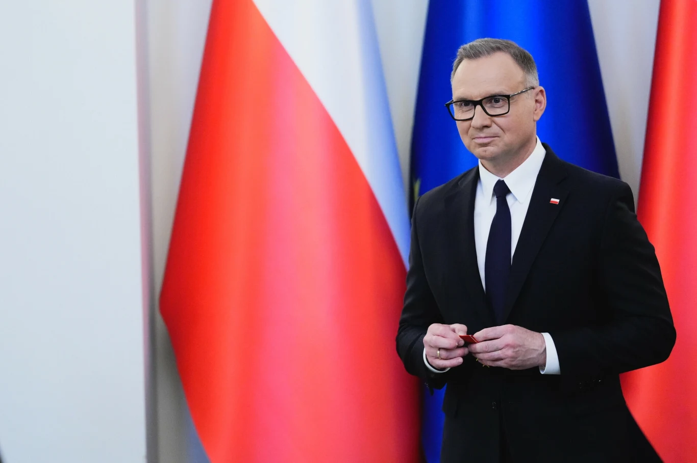 Polsat News: Andrzej Duda nie wróci do pracy na UJ w Krakowie Mężczyzna w okularach ubrany w garnitur stoi na tle polskich flag oraz flag innych krajów europejskich, trzymając niewielki przedmiot w dłoniach.