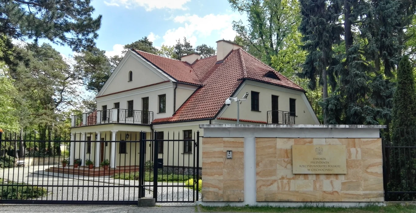 Dworek prezydenta w Ciechocinku Dwukondygnacyjny jasny budynek z czerwonym dachem otoczony zielenią, znajdujący się za czarnym ogrodzeniem. Przed wejściem do obiektu widoczna tablica informacyjna przytwierdzona do ogrodzenia, wskazująca na oficjalny charakter miejsca.