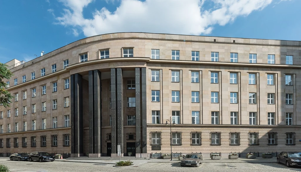 Monumentalny, wielopiętrowy gmach o jasnej elewacji i regularnych rzędach okien, na froncie wyróżniają się wysokie kolumny i wejście główne, przed budynkiem zaparkowane są auta.
