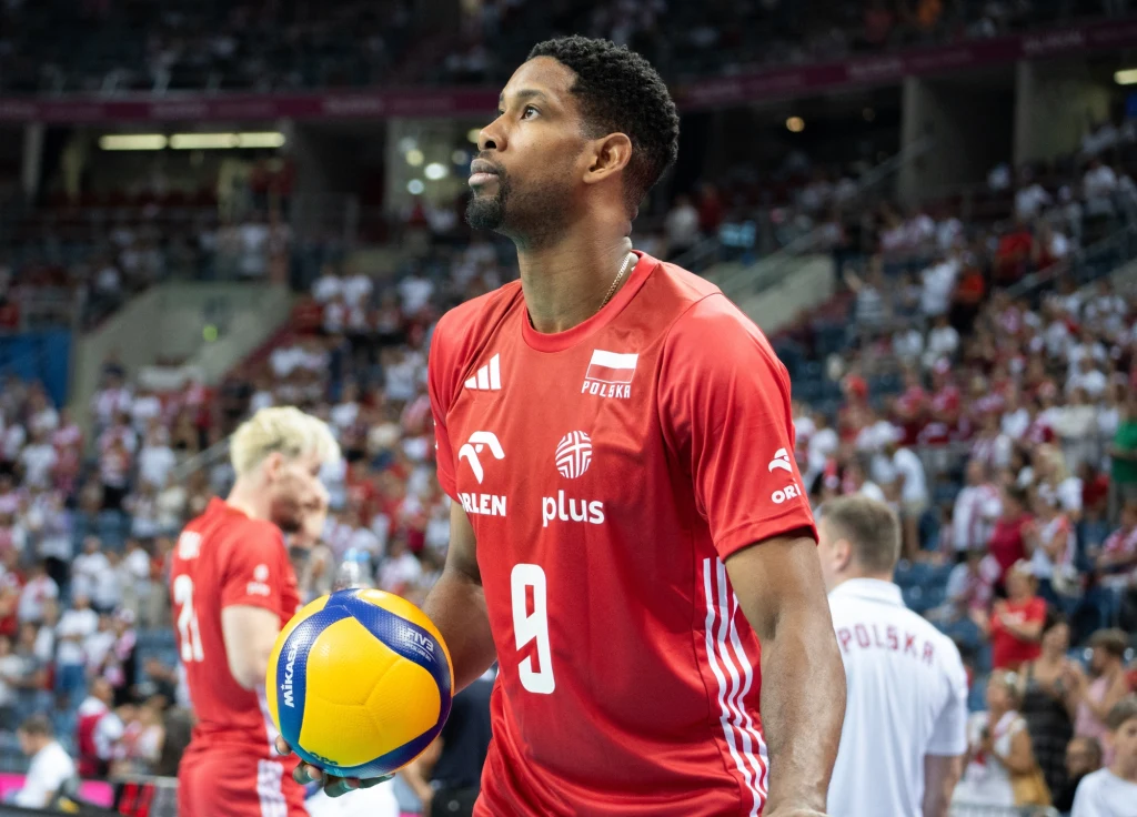 Siatkarz w czerwonym stroju polskiej reprezentacji trzyma piłkę do siatkówki, stojąc na boisku halowym, w tle widoczna pełna kibiców trybuna i inni zawodnicy w czerwonych i białych strojach.
