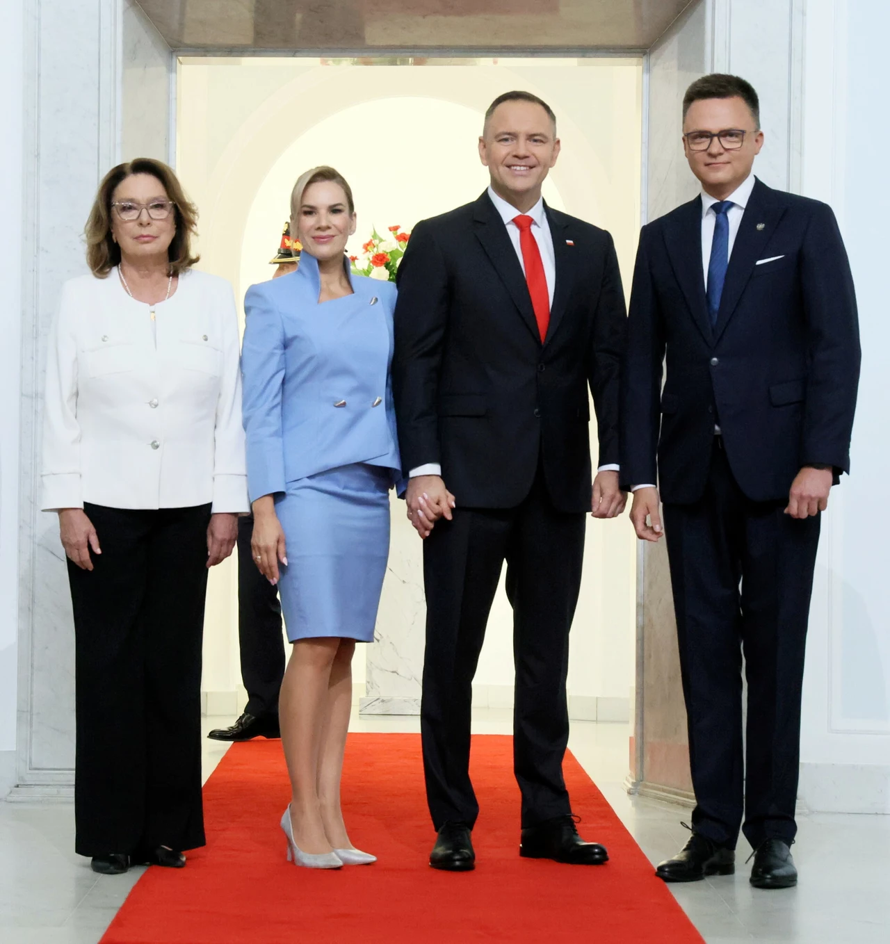 Cztery elegancko ubrane osoby stoją na czerwonym dywanie w formalnym, jasnym wnętrzu, dwie kobiety i dwóch mężczyzn w eleganckich strojach formalnych, wszyscy skierowani do obiektywu, w tle widoczne kwiaty.