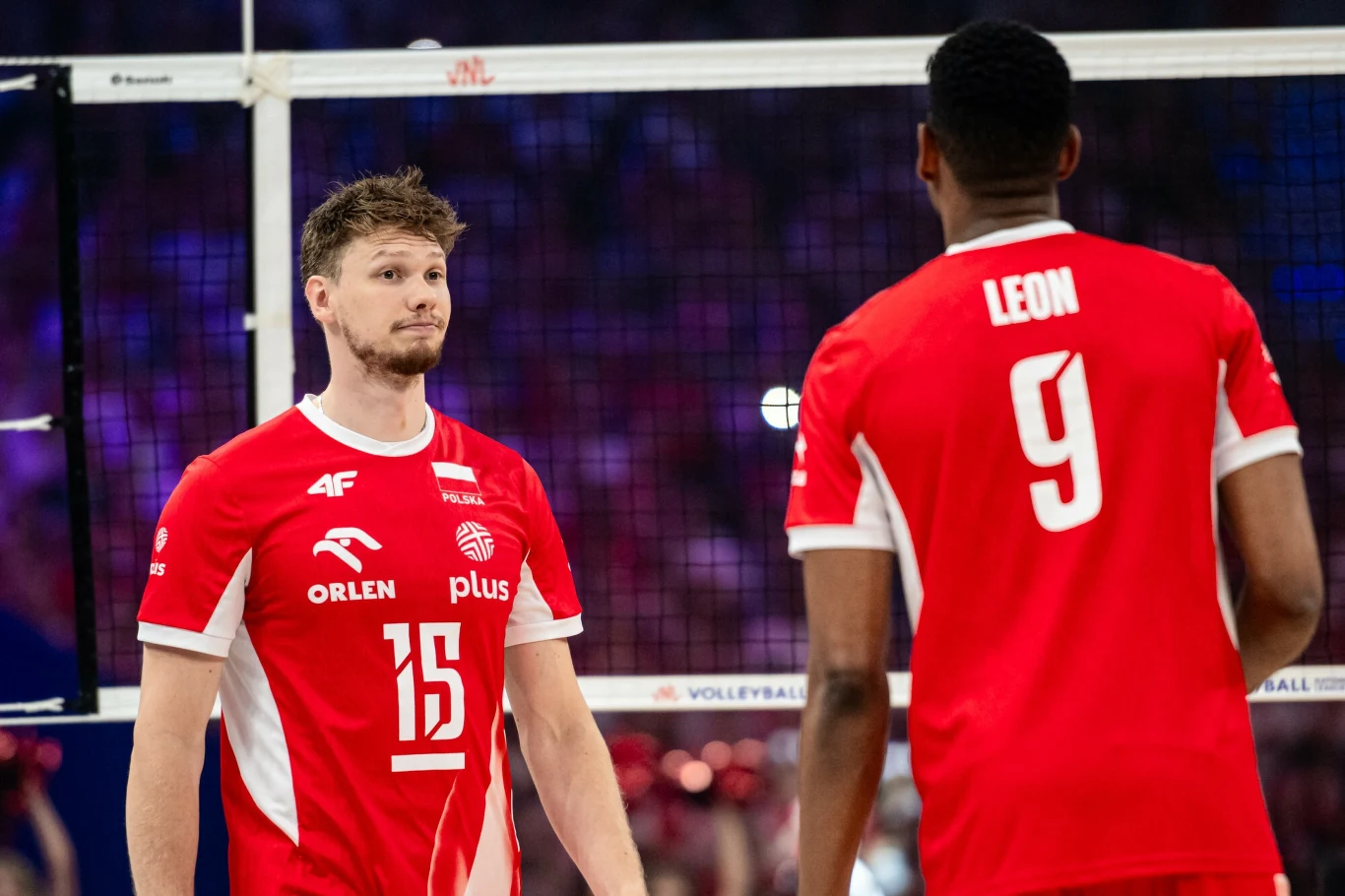 Jakub Kochanowski i Wilfredo Leon Dwóch siatkarzy reprezentacji Polski w czerwonych strojach podczas meczu, jeden z numerem 15 stojący przodem, drugi z numerem 9 z napisem Leon na plecach stojący tyłem, w tle siatka oraz zapełnione trybuny.