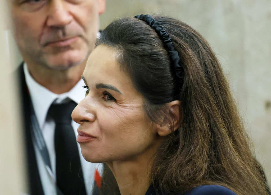Marta Kaczyńska postawiła na naturalność Kobieta z długimi, ciemnymi włosami spiętymi opaską, ubrana w formalny strój, patrzy lekko z boku z subtelnym uśmiechem, w tle widoczny mężczyzna w garniturze i krawacie, rozmyty betonowy kontekst.
