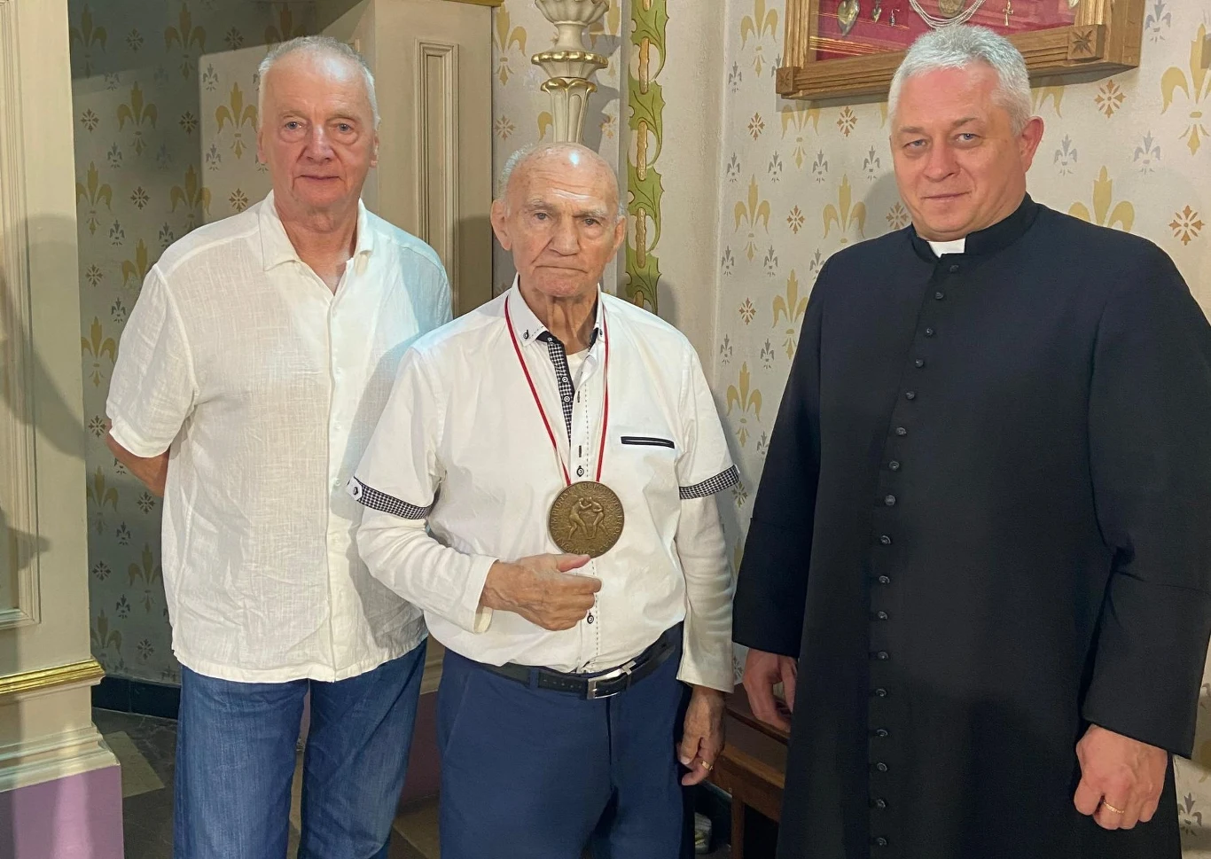 Marian Kasprzyk z proboszczem Andrzejem Zawadą i przyjacielem Józefem Sojką Trzech mężczyzn stoi obok siebie w eleganckim, klasycznie urządzonym pomieszczeniu, środkowy mężczyzna trzyma w dłoni dużą medalę zawieszoną na szyi, po prawej stronie znajduje się ksiądz w czarnej sutannie, pozostali ubrani są w jasne koszule.