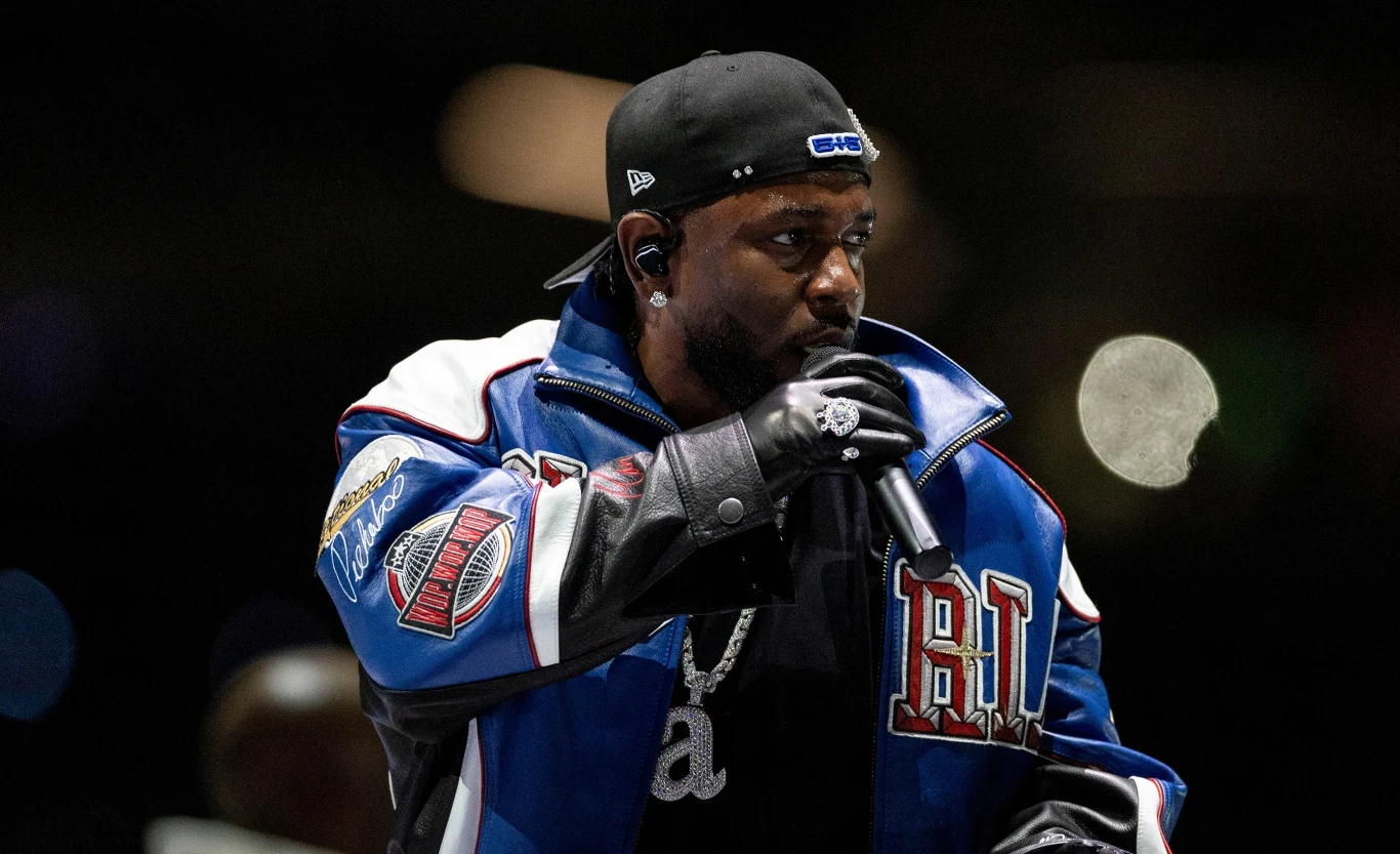 Kendrick Lamar zagra w Warszawie 6 sierpnia 2025 Mężczyzna ubrany w sportową kurtkę i czarną czapkę z daszkiem, trzymający mikrofon i wykonujący utwór na scenie, w tle rozmyte światła.