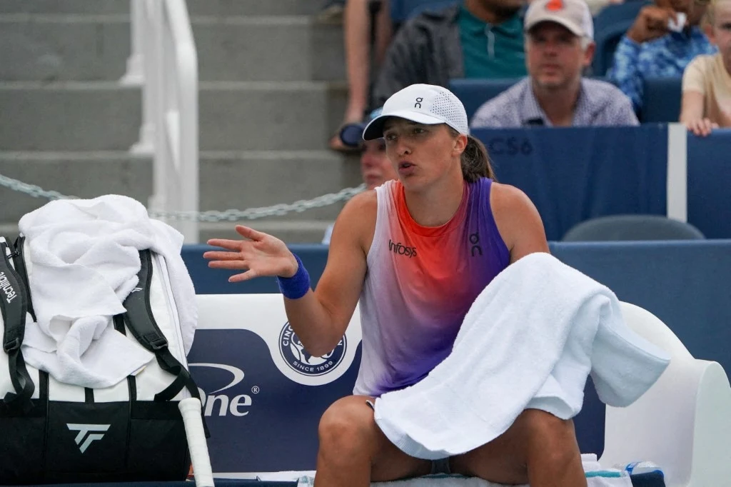 Iga Świątek w WTA 1000 w Cincinnati. Polka przed rokiem odpadła w półfinale Tenisistka siedząca na ławce podczas przerwy w meczu, ubrana w sportowy strój, z czapką na głowie, trzymająca ręcznik i wykonująca gest dłonią, w tle widoczna publiczność oraz torba sportowa.