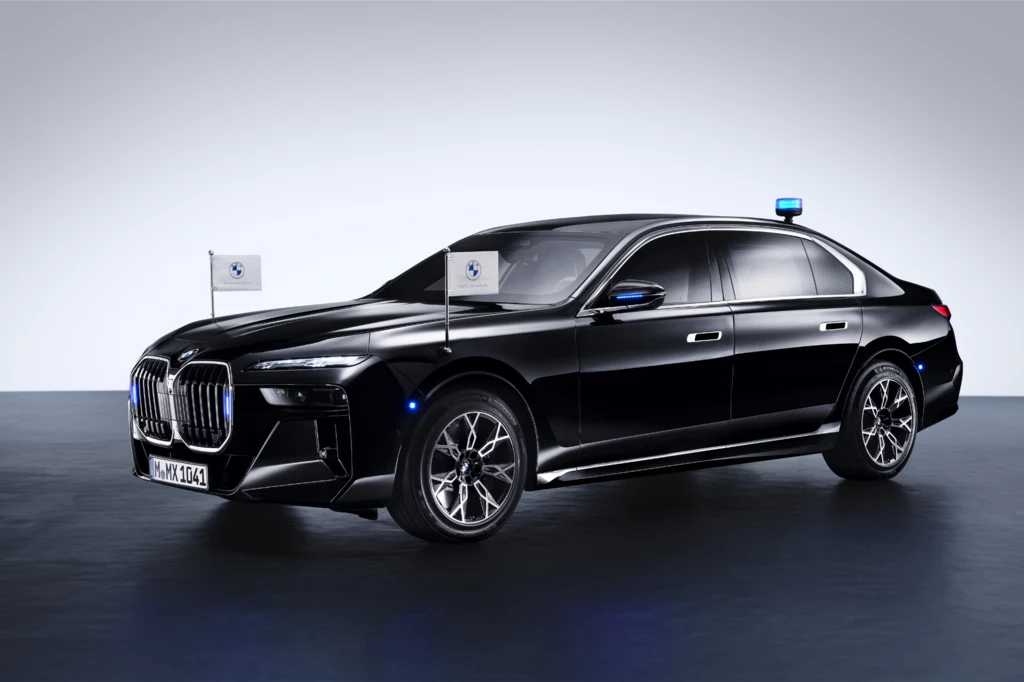 Tak prezentuje się BMW serii 7 w wersji Protection. Czarny luksusowy sedan marki BMW z niebieskim światłem sygnalizacyjnym na dachu oraz dwiema flagami zamocowanymi po bokach maski samochodu.