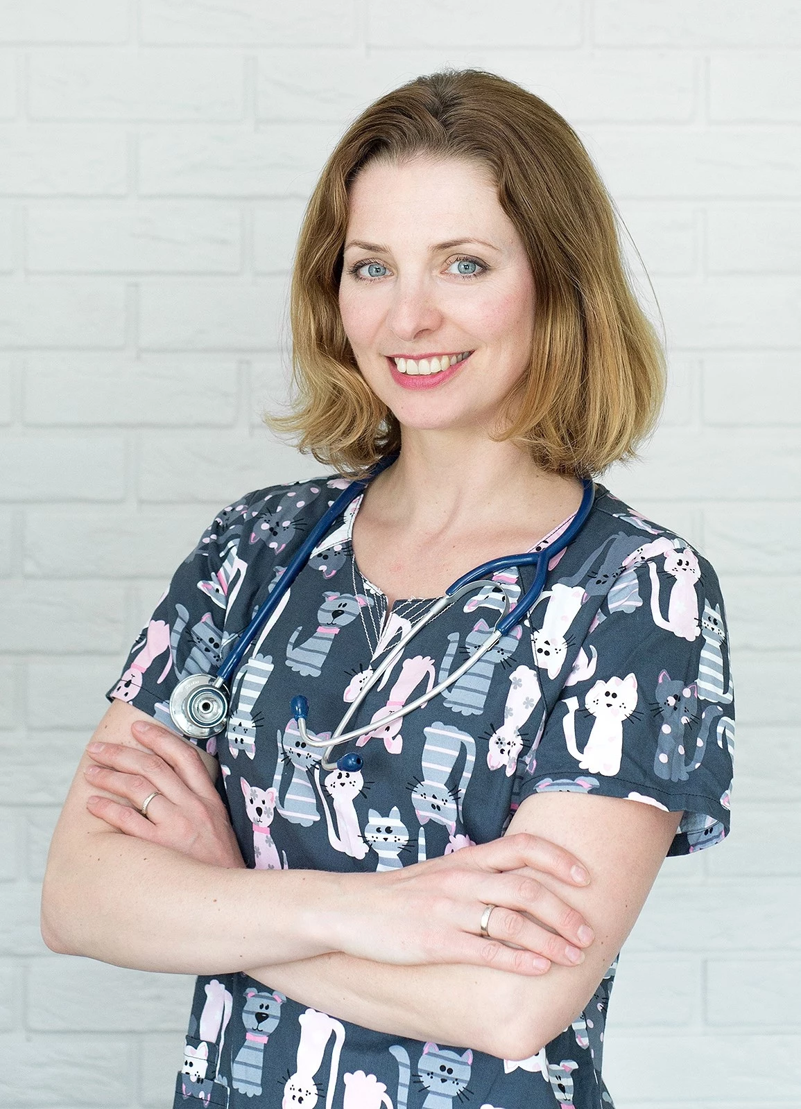 Małgorzata Głowacka, lekarka weterynarii i ekspertka naukowa ds. żywienia zwierząt Kobieta w średnim wieku z jasnymi włosami ubrana w kolorowy fartuch z nadrukiem kotów oraz ze stetoskopem na szyi, stoi z założonymi rękami i uśmiecha się lekko patrząc w stronę aparatu, na tle jasnej ściany z cegły.