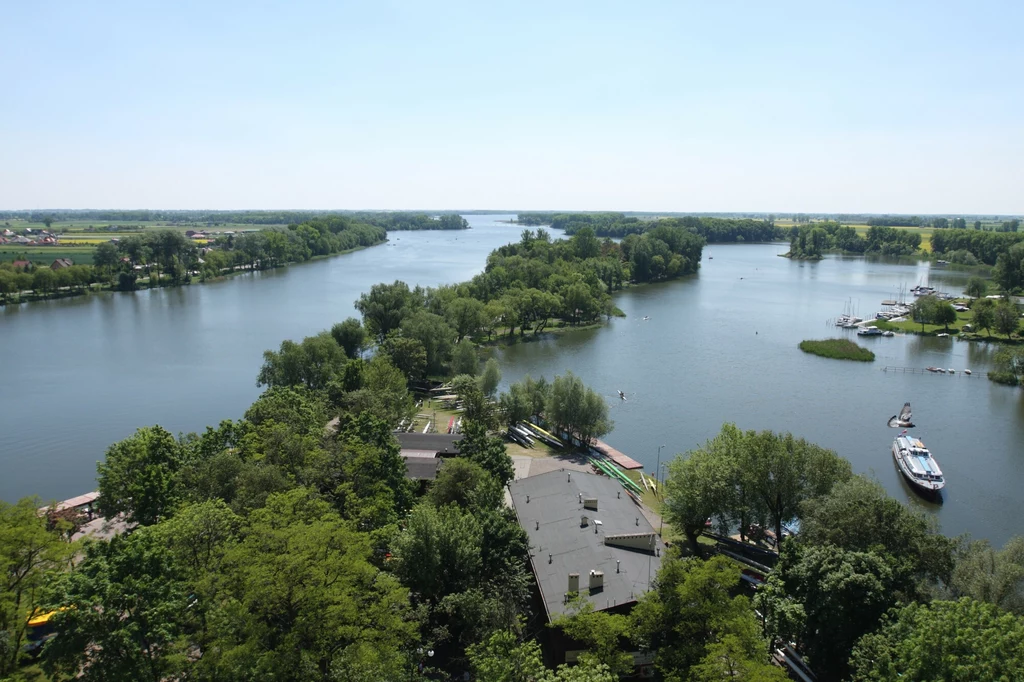 Panorama jeziora Gopło z pasem drzew po obu stronach, na pierwszym planie budynki i przystań z łodzią, otoczenie zieleni oraz szerokie wody rzeki tworzą spokojny krajobraz.