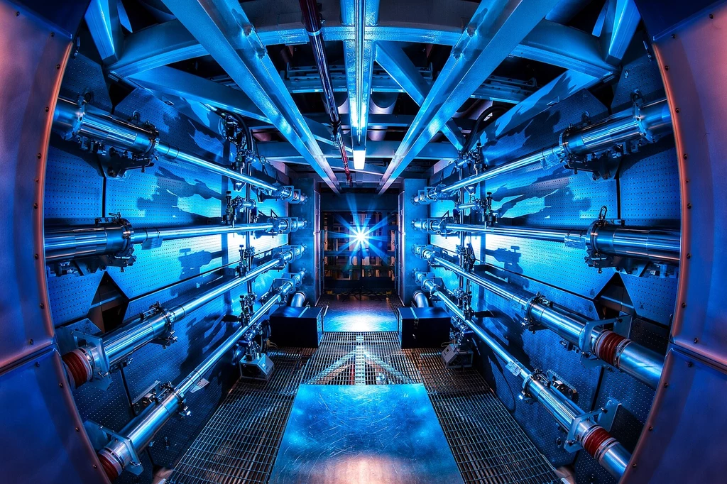 W eksperymentach LLNL energia termojądrowa jest generowana poprzez skierowanie 192 potężnych wiązek laserowych na maleńką kapsułkę paliwową wypełnioną deuterem i trytem, wyzwalając reakcję łańcuchową pod ekstremalnym ciśnieniem i ciepłem Symetryczne wnętrze zaawansowanego technologicznie urządzenia, oświetlone intensywnym niebieskim światłem, z licznymi rurami i metalowymi elementami prowadzącymi do centralnego punktu, w którym widoczny jest jasny świetlny impuls.