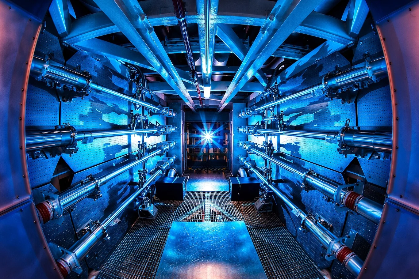 Symetryczne wnętrze zaawansowanego technologicznie urządzenia, oświetlone intensywnym niebieskim światłem, z licznymi rurami i metalowymi elementami prowadzącymi do centralnego punktu, w którym widoczny jest jasny świetlny impuls.
