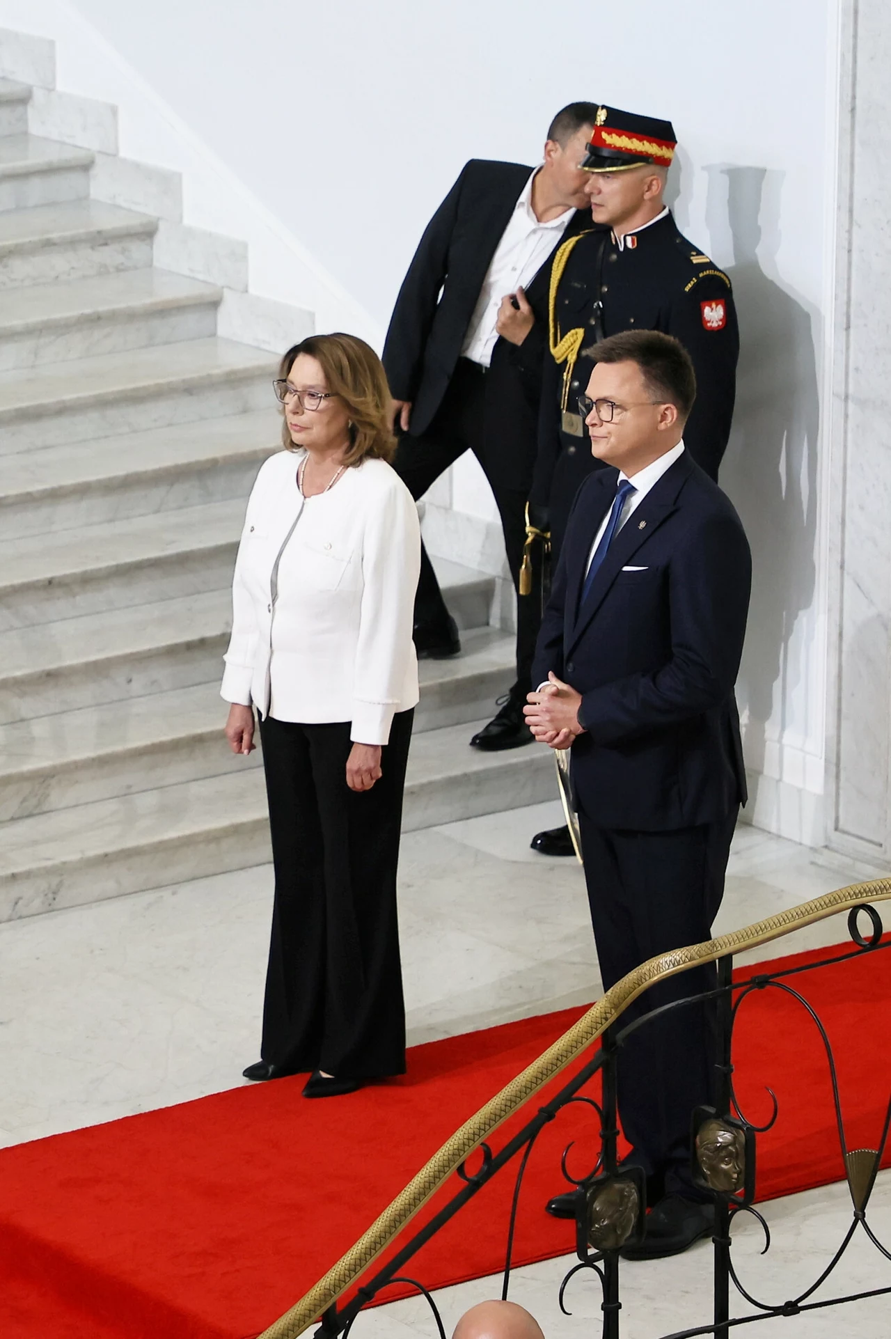 Niektórzy fani mody mogą być zdania, że stylizacja Małgorzaty Kidawy-Błońskiej była zbyt sztampowa, ale może mieć to ukryty cel Dwie elegancko ubrane osoby stoją na czerwonym dywanie, w tle schody z marmuru oraz funkcjonariusze w mundurach pilnujący porządku.