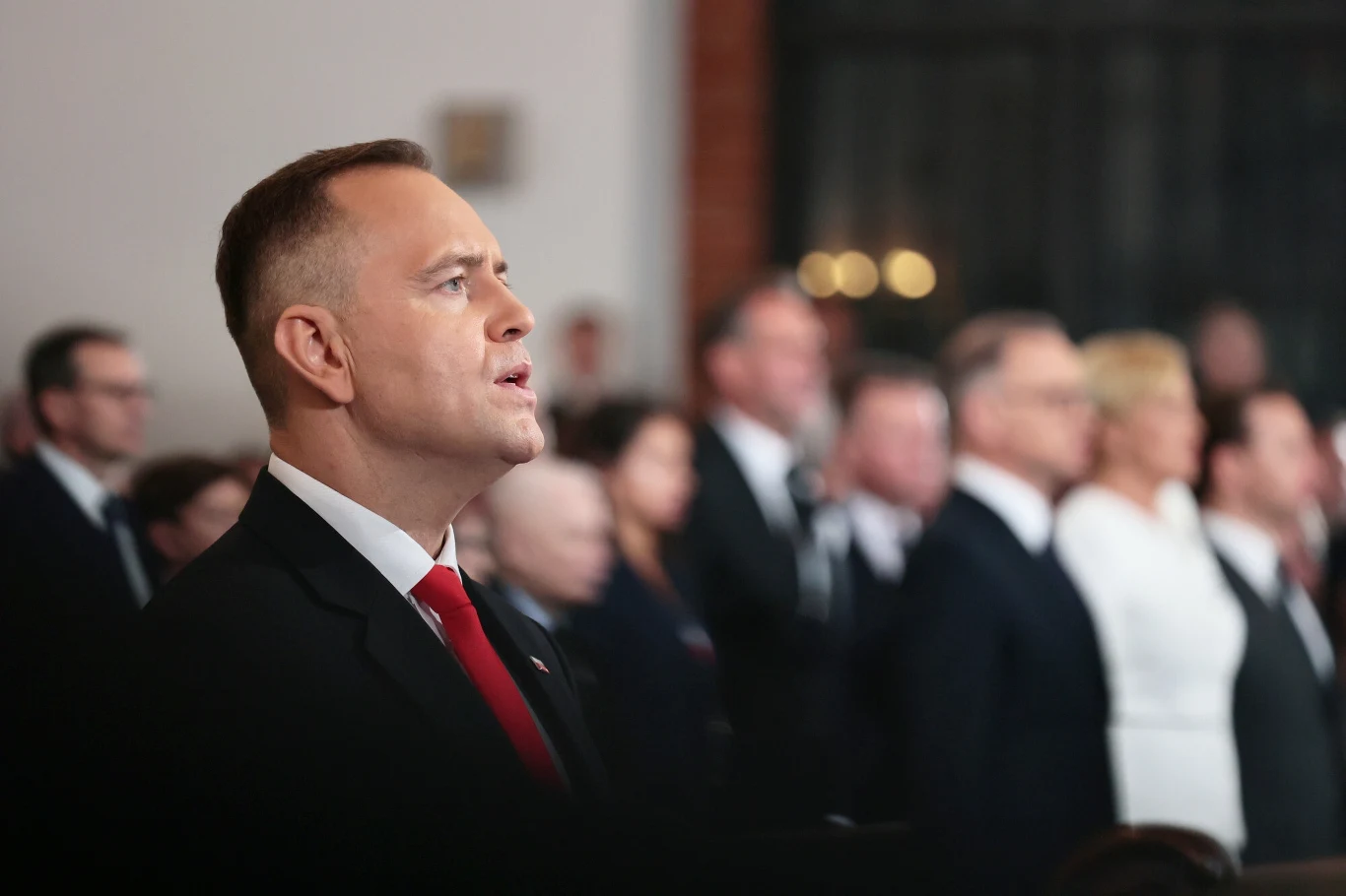 Światowe media odnotowały zaprzysiężenie Karola Nawrockiego Mężczyzna w garniturze i czerwonym krawacie na pierwszym planie śpiewa lub przemawia, w tle widoczna grupa osób w eleganckich strojach, wszyscy stoją w podniosłej atmosferze uroczystości.