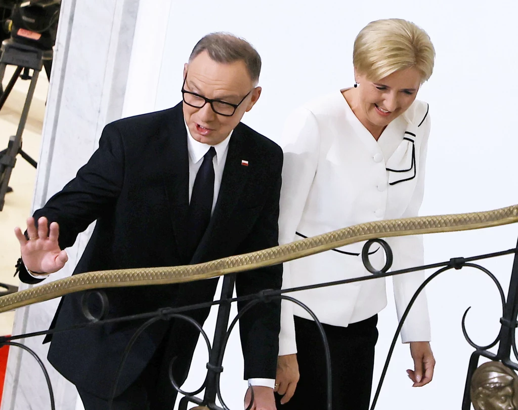 Para elegancko ubranych osób idzie razem po schodach w eleganckim wnętrzu, mężczyzna w garniturze z flagą Polski na klapie, kobieta w białej marynarce z czarnym wzorem, oboje uśmiechnięci.