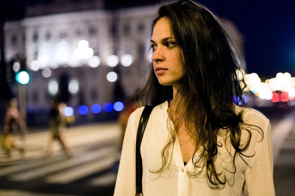 Kobiety czują się realnie zagrożone nawet wieczorem na ulicy Młoda kobieta z długimi, ciemnymi włosami ubrana w jasną bluzkę, stoi na tle rozświetlonego, miejskiego krajobrazu nocą, patrząc w bok z zamyśleniem.