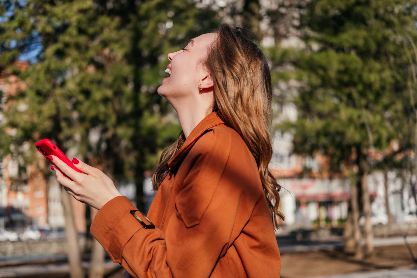Kobieta w rudym płaszczu uśmiecha się szeroko, trzymając w dłoni czerwony smartfon, stojąc na zewnątrz wśród drzew.