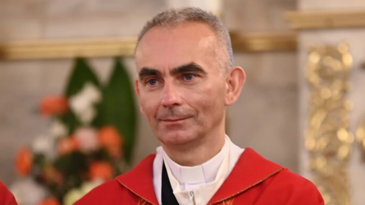 Ks. Krzysztof Dukielski Mężczyzna w średnim wieku o krótkich, siwiejących włosach ubrany w czerwoną szatę liturgiczną, stojący na tle rozmazanego wnętrza kościoła z kwiatami w tle.