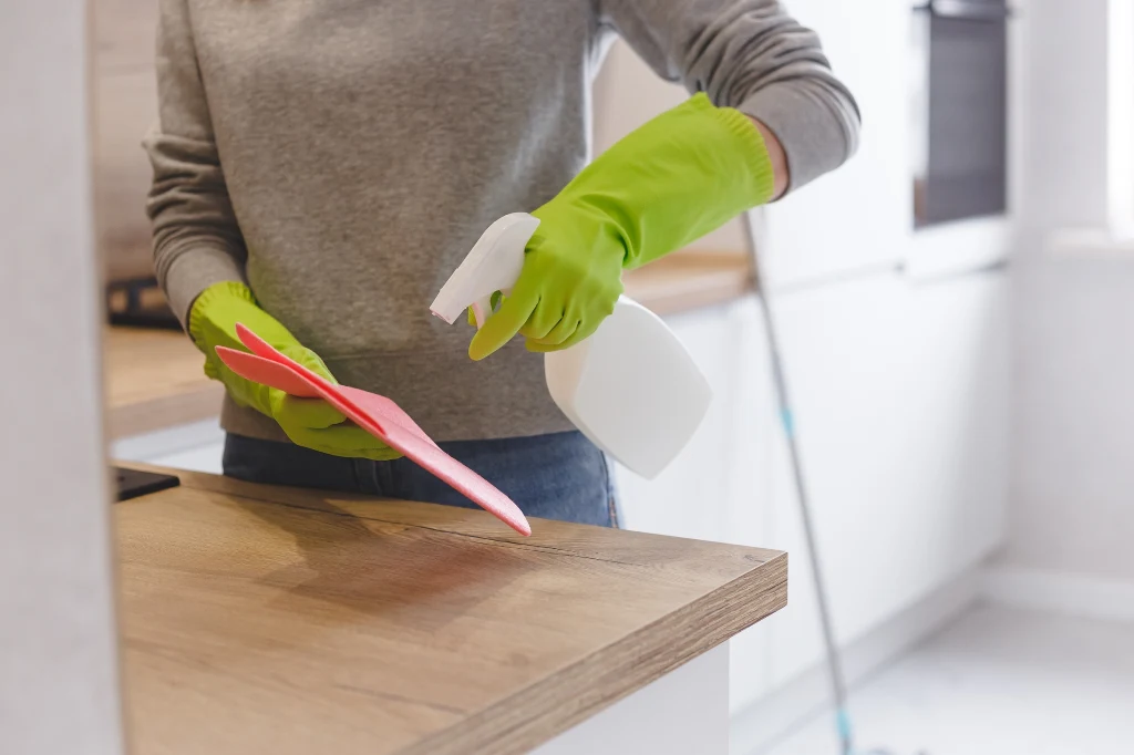 Zbyt mocne detergenty chemiczne mogą doprowadzić do zmatowienia frontów szafek kuchennych Osoba w zielonych rękawiczkach gumowych czyści blat kuchenny przy użyciu ściereczki i butelki ze środkiem czyszczącym, wnętrze kuchni widoczne w tle.
