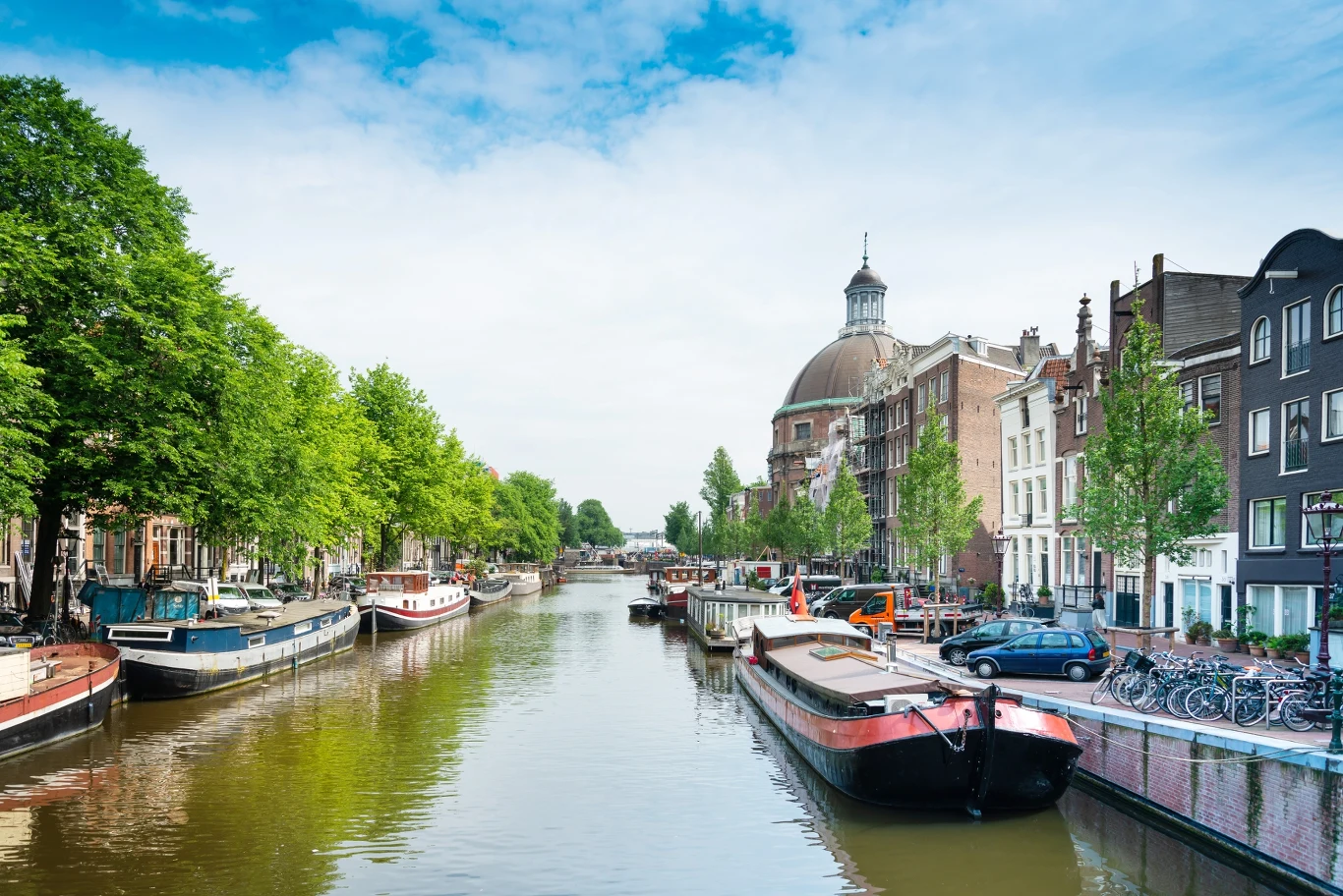 Mieszkańcy Amsterdamu zmęczeni turystami. Stawiają administracji warunki Kanał wodny otoczony przez zacumowane łodzie i barki, wzdłuż którego po obu stronach rosną drzewa oraz stoją rzędy kamienic; po prawej stronie w tle widoczna duża kopuła zabytkowego budynku.