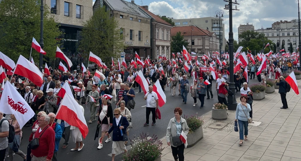 Tłum ludzi maszerujących ulicą, większość uczestników trzyma biało-czerwone flagi Polski, po bokach widać niskie donice z kwiatami, w tle kamienice i niska zabudowa miasta
