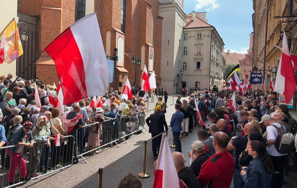 Tłum ludzi zgromadzonych na ulicy z biało-czerwonymi flagami narodowymi Polski, bariery oddzielają uczestników od jezdni, widoczne historyczne kamienice oraz kościół w tle, słoneczna pogoda.
