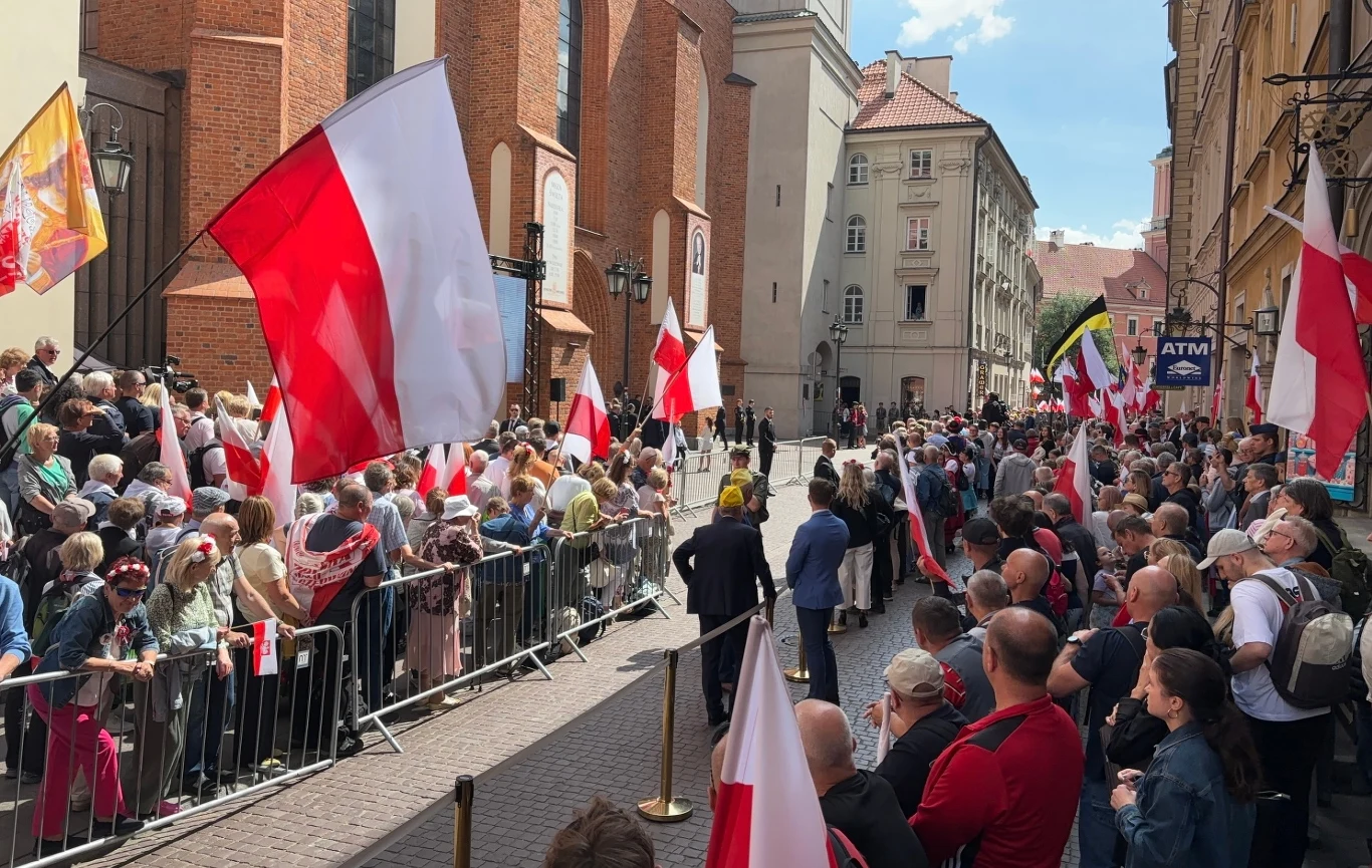 Szczelnie wypełnione ulice wokół warszawskiej archikatedry Tłum ludzi zgromadzonych na ulicy z biało-czerwonymi flagami narodowymi Polski, bariery oddzielają uczestników od jezdni, widoczne historyczne kamienice oraz kościół w tle, słoneczna pogoda.