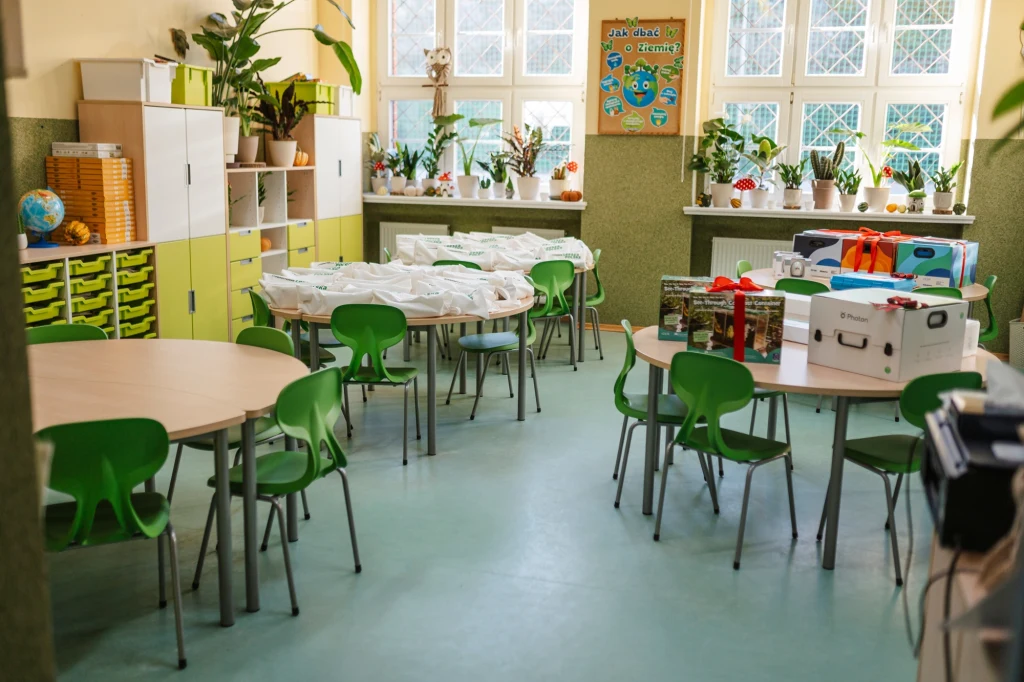 Ekopracownia w SP nr 2 w Jaworze - Fot. UM. Jawor Kolorowa sala lekcyjna z okrągłymi stolikami i zielonymi krzesłami, na których ustawione są różne pudełka i materiały edukacyjne, liczne doniczki z roślinami na parapetach, duże okna i dekoracyjne elementy przy ścianach.