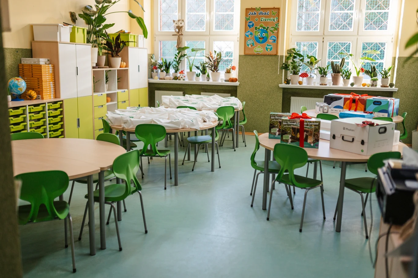 Kolorowa sala lekcyjna z okrągłymi stolikami i zielonymi krzesłami, na których ustawione są różne pudełka i materiały edukacyjne, liczne doniczki z roślinami na parapetach, duże okna i dekoracyjne elementy przy ścianach.