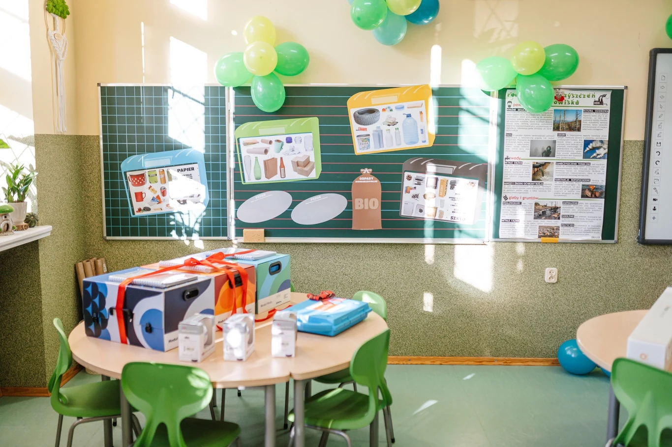 Ustawione przy okrągłym stole zielone krzesła, na stole kolorowe pudełka z prezentami i kartonami, w tle dekoracje z balonów, tablica z plakatami i ilustracjami dotyczącymi segregacji odpadów oraz artykułów ekologicznych.