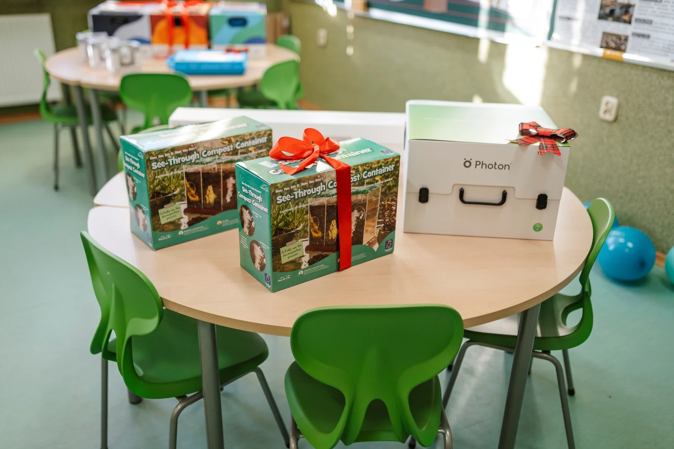 Na okrągłym stole znajdują się dwa pudełka z zestawem edukacyjnym Solo Through the Campfire ozdobione czerwonymi kokardkami oraz duża biała walizka marki Photon, wokół rozstawione zielone krzesła dziecięce.