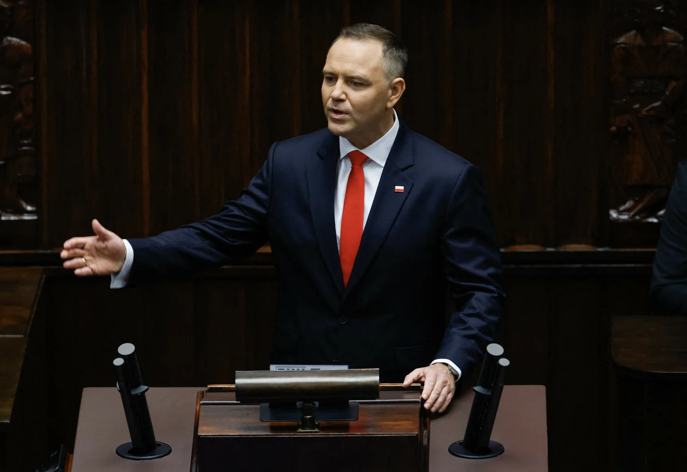 Karol Nawrocki zaprzysiężony na prezydenta Mężczyzna w garniturze z przypiętą flagą Polski, stojący za mównicą sejmową i wykonujący gest ręką w trakcie przemówienia, ciemne wnętrze sali.