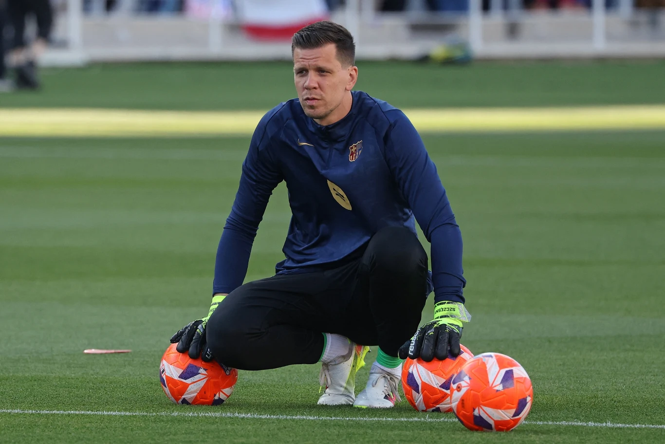 Wojciech Szczęsny Piłkarz w stroju bramkarskim klęczący na murawie boiska, trzymający trzy piłki do piłki nożnej, ujęcie z treningu lub rozgrzewki.