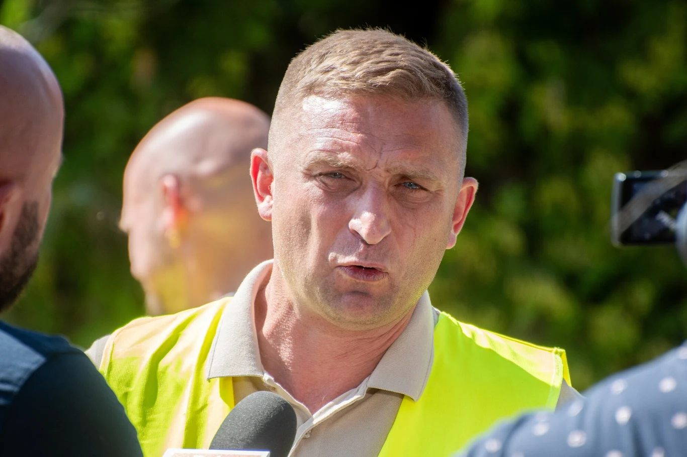 Robert Bąkiewicz ma usłyszeć zarzuty Mężczyzna w kamizelce odblaskowej udzielający wywiadu mediom na zewnątrz, w tle niewyraźne sylwetki innych osób, wokół zielone otoczenie.