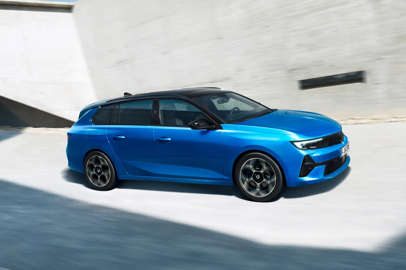 Nowoczesny niebieski samochód kombi jedzie wzdłuż betonowej ściany, wyróżnia się sportową sylwetką, ciemnym dachem i dużymi felgami.