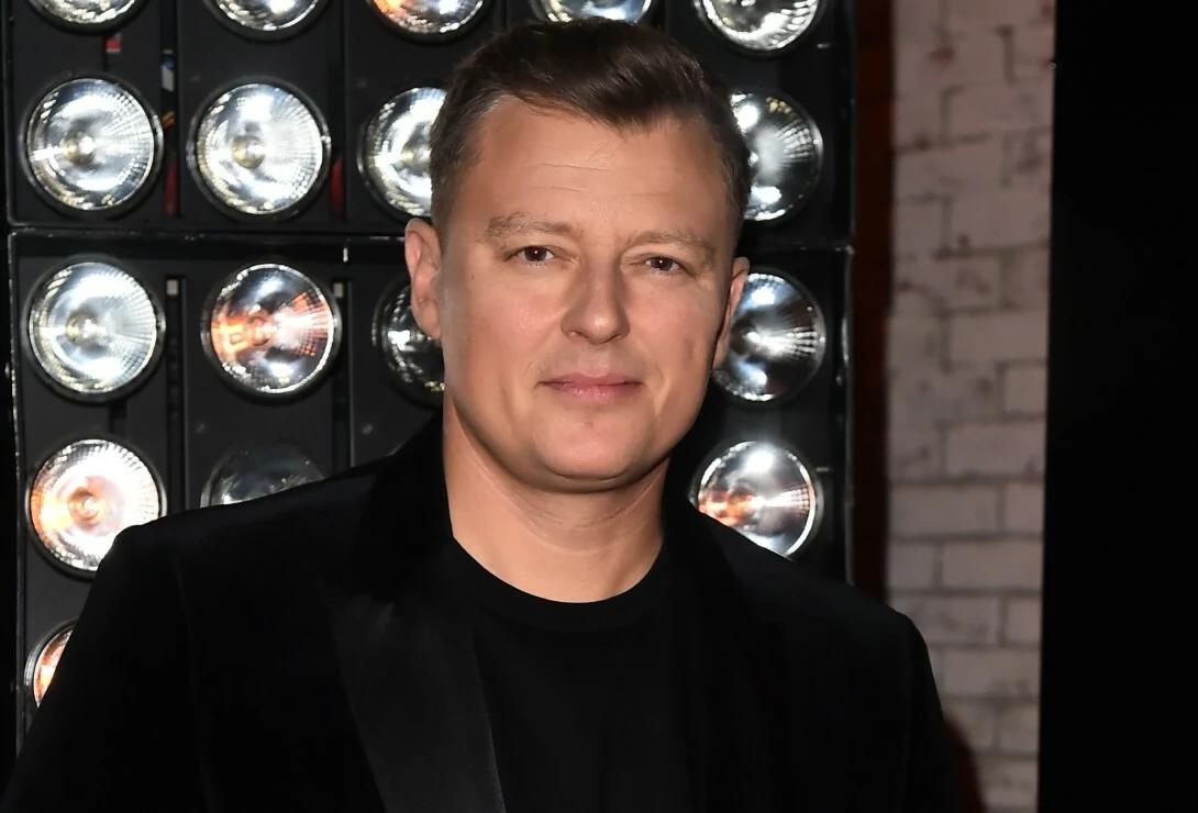 Rafał Brzozowski Mężczyzna w czarnym garniturze stoi na tle ściany z reflektorami i ceglaną ścianą, patrzy prosto w obiektyw z delikatnym uśmiechem.