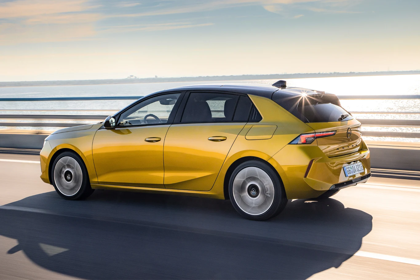 Żółty samochód osobowy typu hatchback jadący mostem w słoneczny dzień, z widokiem na wodę w tle.