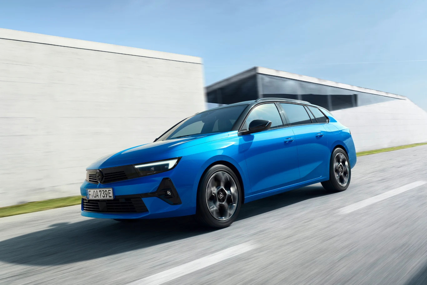 Jasnoniebieski samochód osobowy najnowszej generacji porusza się po jasnej asfaltowej drodze na tle nowoczesnego budynku o minimalistycznej architekturze.