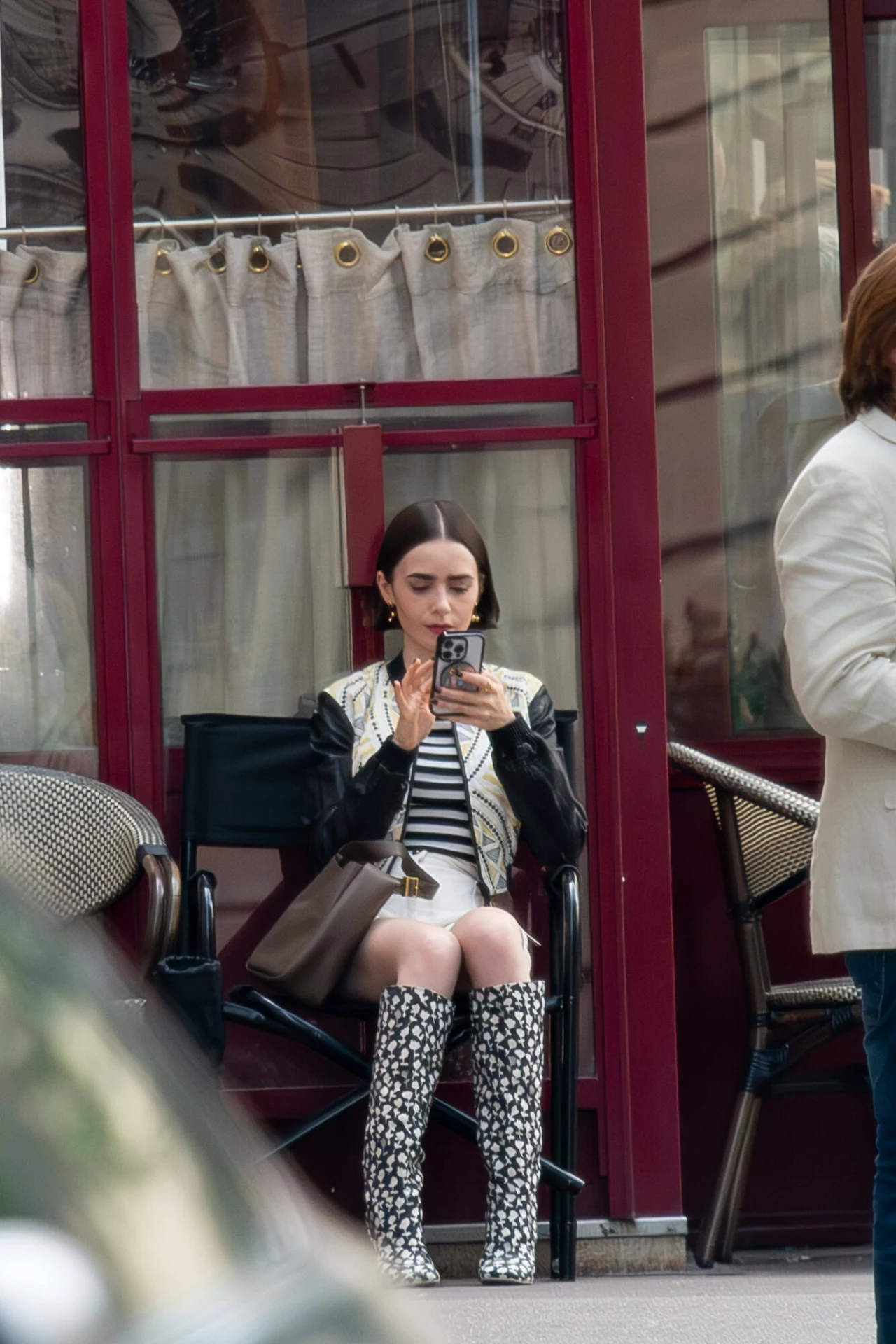Kobieta siedząca na krześle przed lokalem, ubrana w czarno-białą stylizację z długimi wzorzystymi kozakami, trzyma telefon i skupia się na jego ekranie, obok znajduje się brązowa torebka.
