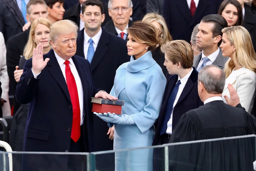 Melania Trump w swojej kultowej już dziś kreacji Mężczyzna w ciemnym płaszczu i czerwonym krawacie podnosi prawą rękę do przysięgi, stojąc przed grupą osób, obok kobieta w jasnoniebieskim płaszczu trzyma otwartą księgę, a wokół nich zgromadzeni są ludzie w oficjalnych strojach, ceremonia odbywa się n...