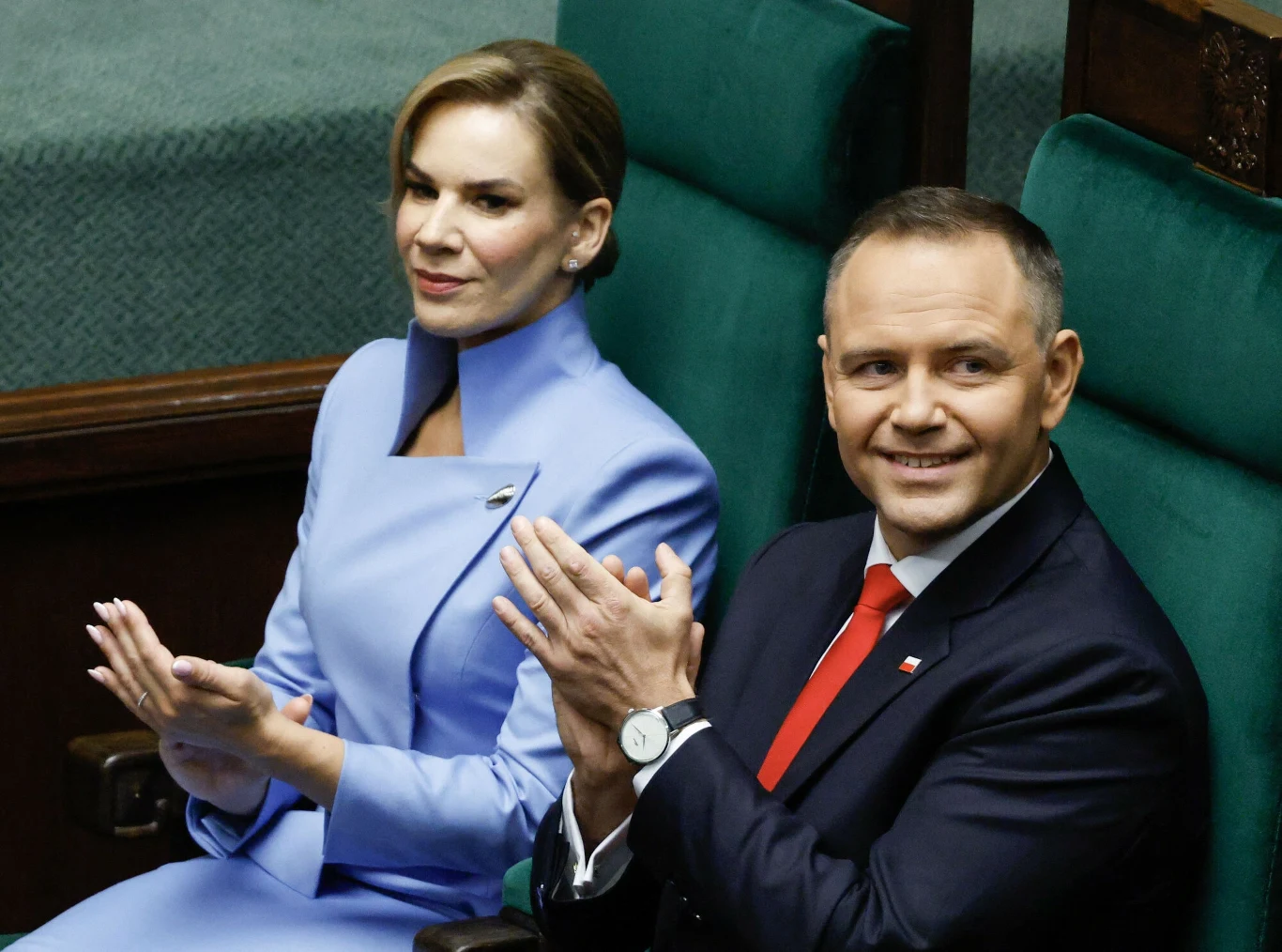 Dwóch elegancko ubranych ludzi siedzących obok siebie na zielonych fotelach, kobieta ubrana w jasnoniebieski kostium, mężczyzna w garniturze z czerwoną krawatą, oboje uśmiechnięci i bijący brawo.