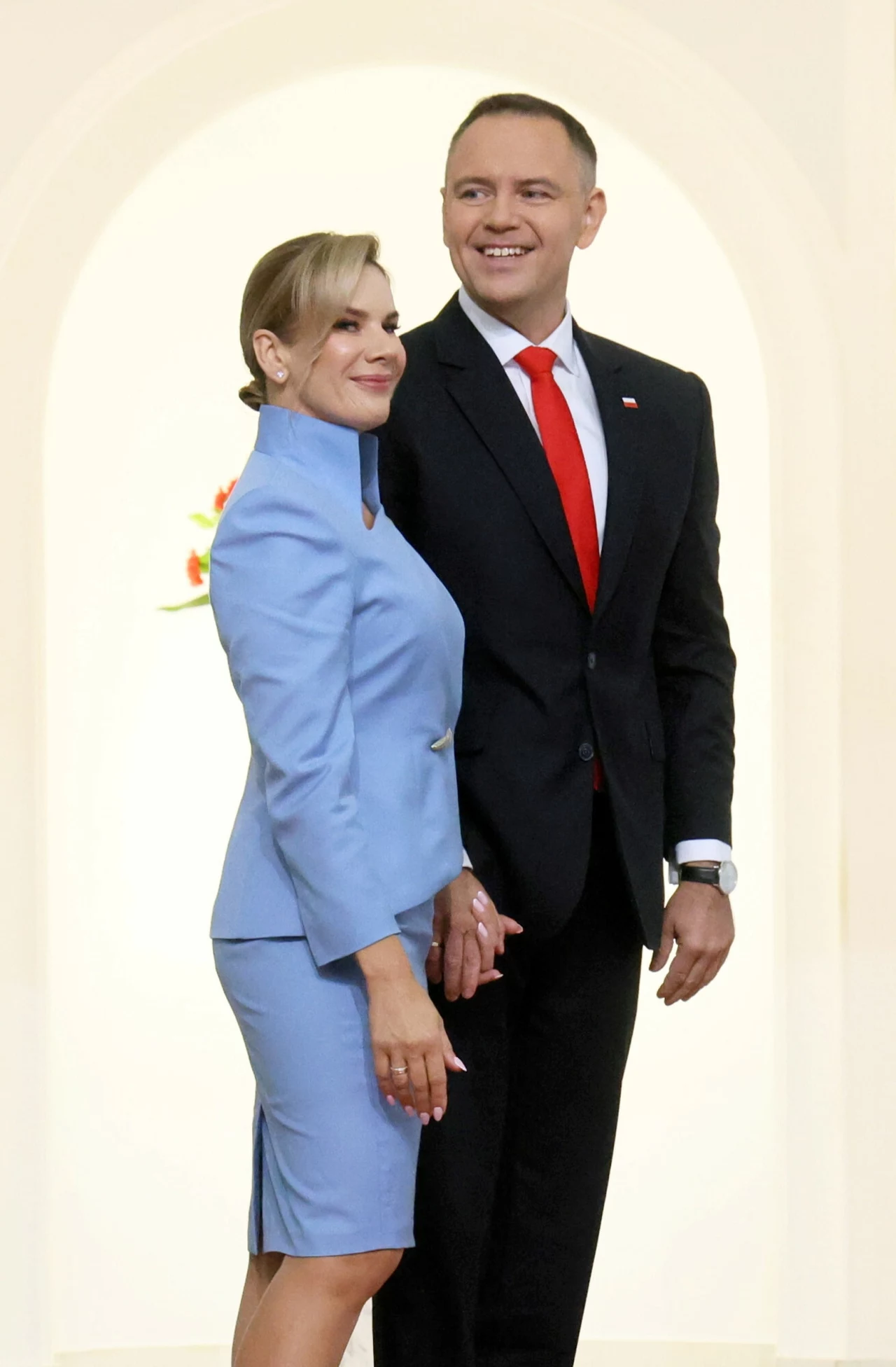 Marta Nawrocka z mężem Elegancka para uśmiechniętych osób w oficjalnych strojach, kobieta w niebieskim kostiumie, mężczyzna w czarnym garniturze z czerwonym krawatem, jasne tło podkreślające sylwetki.